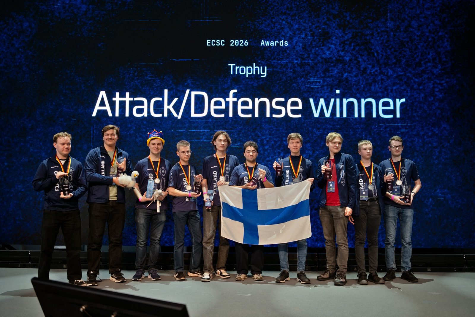 The Finnish team with flag, trophies and medals on stage in front of the screen reading “Attack/Defense winner”.