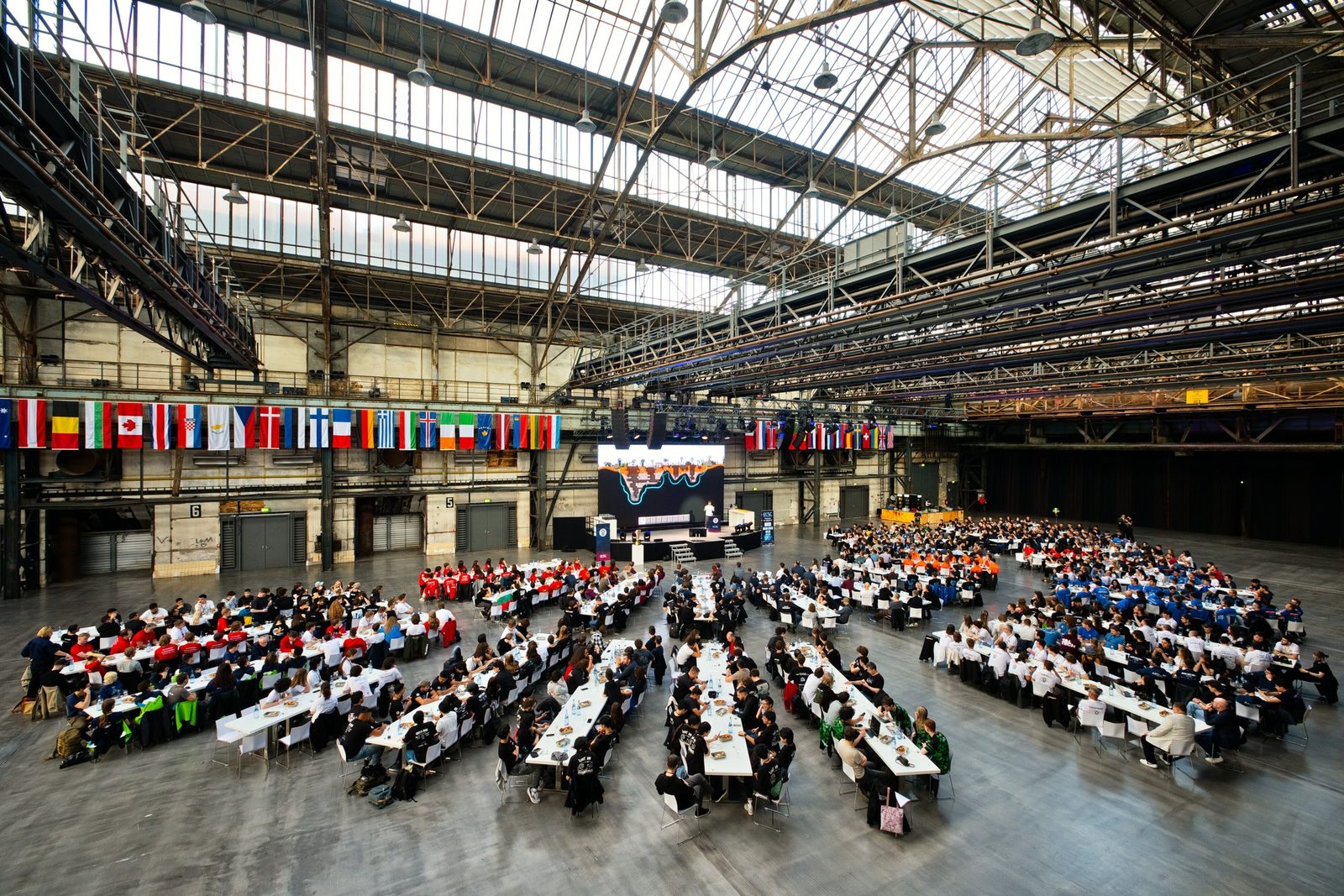 View from above into Jahrhunderthalle Bochum: hundreds of participants at long tables, flags and the steel and glass roof above.