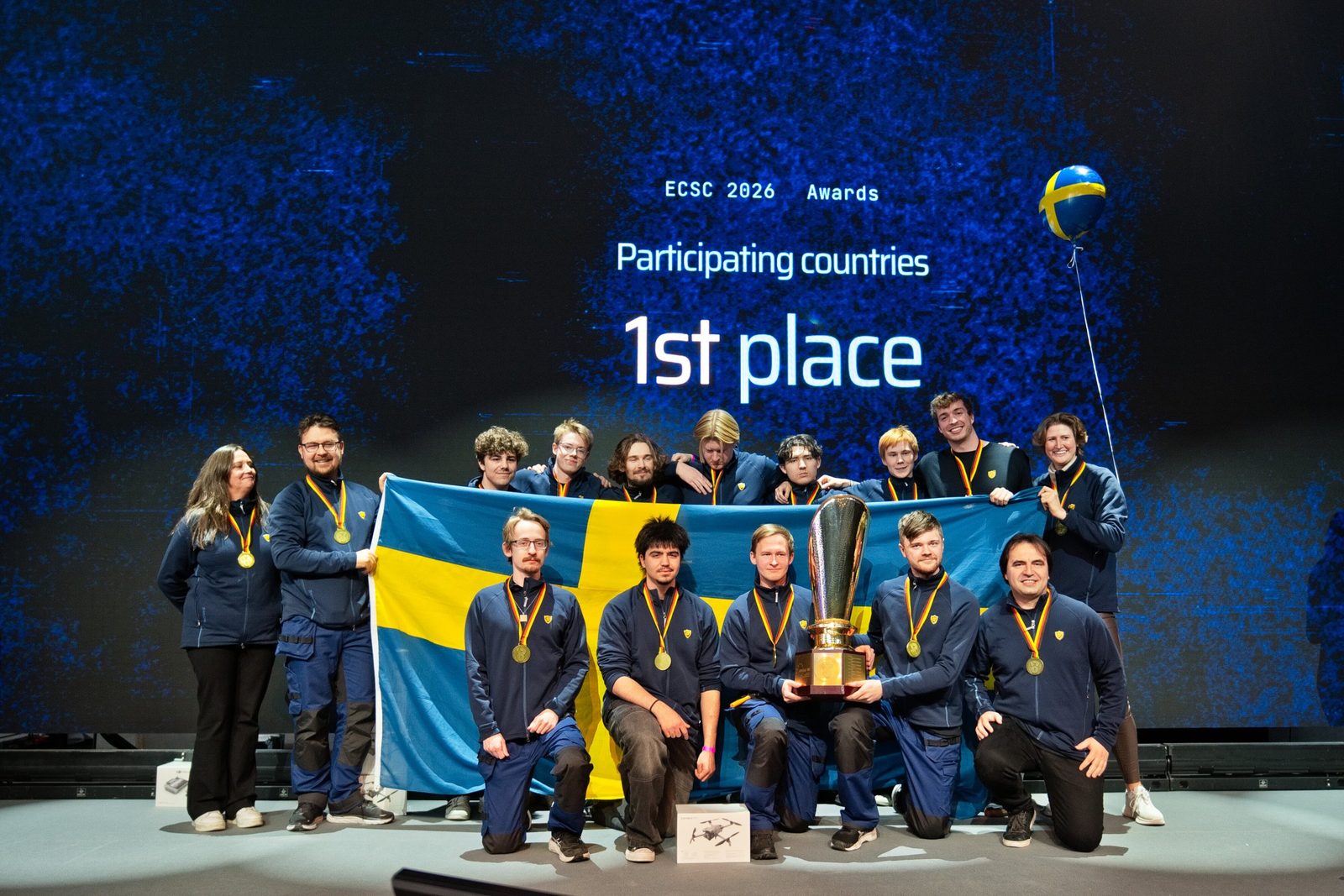 The Swedish team with flag, trophy and gold medals on stage in front of the screen reading “1st place”.
